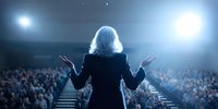 Woman with white hair in black suit addressing large audience from stage with dramatic lighting, perfect for business presentation or leadership concept.