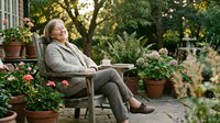 Senior woman relaxing in cozy garden chair surrounded by blooming flowers and potted plants on sunny patio for peaceful retirement lifestyle.