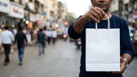 Middle-aged man of South Asian descent wearing dark blue shirt holding blank white shopping bag on busy urban street with pedestrians and storefronts in background.