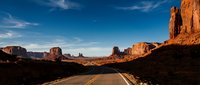 Empty desert highway stretching toward iconic red rock formations under clear blue sky, suitable for travel, adventure, and road trip concepts.