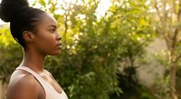 Young woman with dark skin tone and black hair in a bun, wearing a light pink sleeveless top, shown in profile outdoors among green foliage during golden hour lighting.