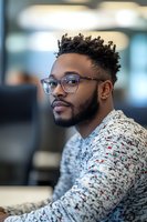 Young African American professional in glasses and patterned sweater looking thoughtfully aside while working in modern office environment. Soft focus portrait captures natural lighting.