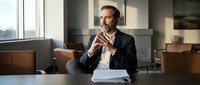 Professional businessman in suit sitting at conference table during meeting, thoughtful pose with hands clasped, modern office interior with natural lighting.