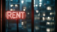 Glowing red neon rent sign in window at night with blurred city lights and apartment buildings in background creates moody urban atmosphere.