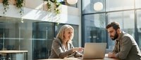 Professional blonde businesswoman and bearded male colleague collaborating on laptop in modern office with natural lighting and glass architecture.