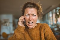 Frustrated middle-aged Caucasian woman yelling during phone call wearing orange sweater in modern office expressing anger and stress at work.