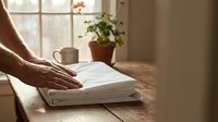 Hands placing neatly folded white linen on a rustic wooden table by a sunlit window with a potted plant and mug in the background.