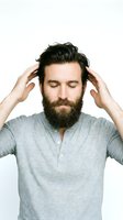 Man with full beard and dark hair touching head with both hands, wearing gray henley shirt against white background for grooming and lifestyle content.