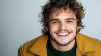 Young Caucasian man with curly brown hair and beard wearing mustard yellow jacket and green sweater smiling genuinely at camera against light gray background.