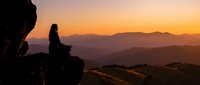Woman silhouette meditating on mountain cliff during golden sunset with layered mountain ranges in background creating peaceful spiritual moment.