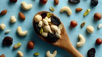 Mixed nuts and dried fruits in wooden spoon on blue background. Healthy snacking, trail mix ingredients including cashews, almonds, hazelnuts, raisins.