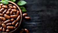 Fresh roasted coffee beans in wooden bowl with scattered beans and green leaves on dark rustic background, dramatic lighting for food photography.