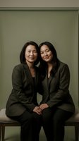 Two Asian women, mature and young, in matching dark blazers sit together smiling against a muted green studio backdrop.