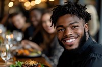 Young African American man with beard smiling at dinner table with friends, warm bokeh lighting, casual gathering, social dining event.