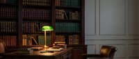 Classic home office interior with wooden desk, green banker lamp, leather chair and floor-to-ceiling bookshelves filled with vintage books in warm lighting.