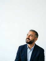 Middle-aged man with salt-and-pepper beard and dark hair wearing navy blazer over light blue shirt, looking upward thoughtfully against white background. Isolated portrait for corporate and business use.