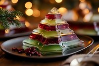 Colorful layered jelly dessert in pyramid shape on dark plate with festive bokeh lights and pine branch in background, holiday table setting.