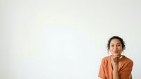 Young man with curly dark hair and olive complexion wearing rust-colored shirt, hand near face in thoughtful pose against white background, suitable for business, lifestyle, and editorial design projects.