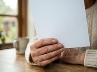 Senior man with weathered hands holding blank white paper on wooden table indoors, suitable for business communications, healthcare messaging, legal documents, or retirement planning concepts.