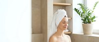 Young woman relaxing in modern bathroom after shower with towel wrapped around head, enjoying peaceful spa moment at home with natural lighting.