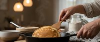 Young adult male with light skin tone preparing fresh baked goods in rustic kitchen setting, pouring flour or powdered ingredient over pastry in cast iron skillet, warm ambient lighting.