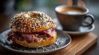 Gourmet breakfast bagel with crispy bacon and melted cheese on seeded bun served alongside steaming coffee cup on wooden table for morning meal concepts.