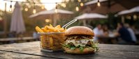 Gourmet burger with fresh lettuce and fries in metal basket on wooden table at outdoor restaurant patio with string lights and umbrellas.