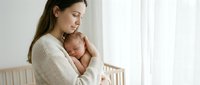 Young Caucasian mother in cream sweater tenderly holding sleeping newborn baby in bright nursery room with natural window light for parenting content.