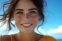 Young woman with bright smile and natural freckles wearing yellow top against blue summer sky, windswept hair and glowing skin capture carefree joy and wellness.