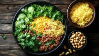 Fresh bowl with minced meat, yellow rice, cucumber slices, green herbs and peanuts served on rustic wooden table, healthy lunch concept.