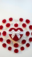 Red heart-shaped gift box with white bow surrounded by rose petals and baby's breath on white background, romantic Valentine's Day concept.