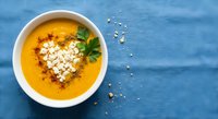 Creamy pumpkin soup with heart-shaped feta cheese and herbs in white bowl on blue wooden background, healthy comfort food for autumn dining.