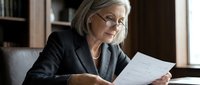 Senior woman with gray hair and glasses wearing dark blazer reviewing documents at desk in office setting.