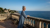 Senior Caucasian woman in blue dress standing on Mediterranean coastal terrace balcony overlooking azure sea and historic architecture during golden hour.