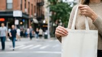 Woman holding blank canvas tote bag on urban street with pedestrians and brick buildings in background, suitable for branding, marketing, and retail mockup purposes.