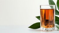 Glass of amber-colored tea or whiskey with green leaves on white background, minimalist composition for food and beverage marketing.