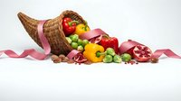 Wicker cornucopia overflowing with fresh vegetables including red and yellow bell peppers, Brussels sprouts, and pomegranate with pink ribbon on white background.