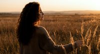 Young woman with curly hair in knit sweater standing in golden wheat field at sunset, peaceful countryside moment for wellness and nature connection.