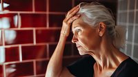 Senior Caucasian woman with worried expression against red brick wall, dramatic side lighting highlighting facial features and silver hair.