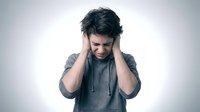 Young man in gray hoodie holding head in hands showing stress, headache, depression, mental health struggles, emotional pain and overwhelm.