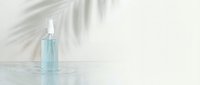 Transparent glass spray bottle with light blue liquid on white surface with palm leaf shadow, isolated for cosmetics and skincare packaging design.