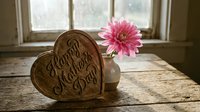 Wooden heart with Happy Mother's Day text and pink gerbera flower in rustic vase on weathered table by window for greeting card design.