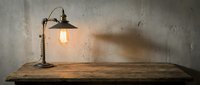 Vintage desk lamp with warm filament bulb casting shadow on rustic wooden table against weathered concrete wall, industrial interior design element.