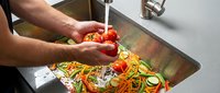 Adult male hands washing fresh red bell peppers and sliced vegetables in stainless steel sink for food preparation and cooking.
