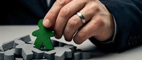 Close-up of male hand placing green wooden figure among gray pieces on white surface, symbolizing uniqueness, leadership, and strategic business decision-making.