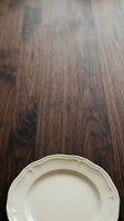 Close-up of cream-colored decorative plate with scalloped rim on dark wood grain surface. Overhead view for tableware, dining, and interior design projects.
