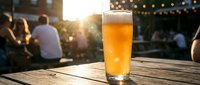 Fresh beer glass on wooden table at outdoor restaurant terrace during golden hour with blurred people socializing in warm evening atmosphere.