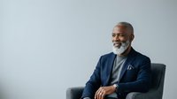 Mature African American male professional wearing navy blazer and light blue shirt, seated in gray chair against white background, smiling with confident expression, suitable for corporate, business, and leadership concepts.
