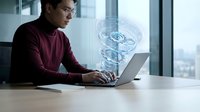 Young Asian male professional working on laptop with holographic DNA helix projection in modern office environment for biotechnology and genetic research applications.