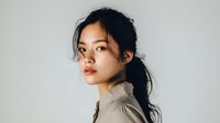 Young Asian woman with dark hair in ponytail against neutral gray background, wearing light-colored high collar top, looking pensively at camera.